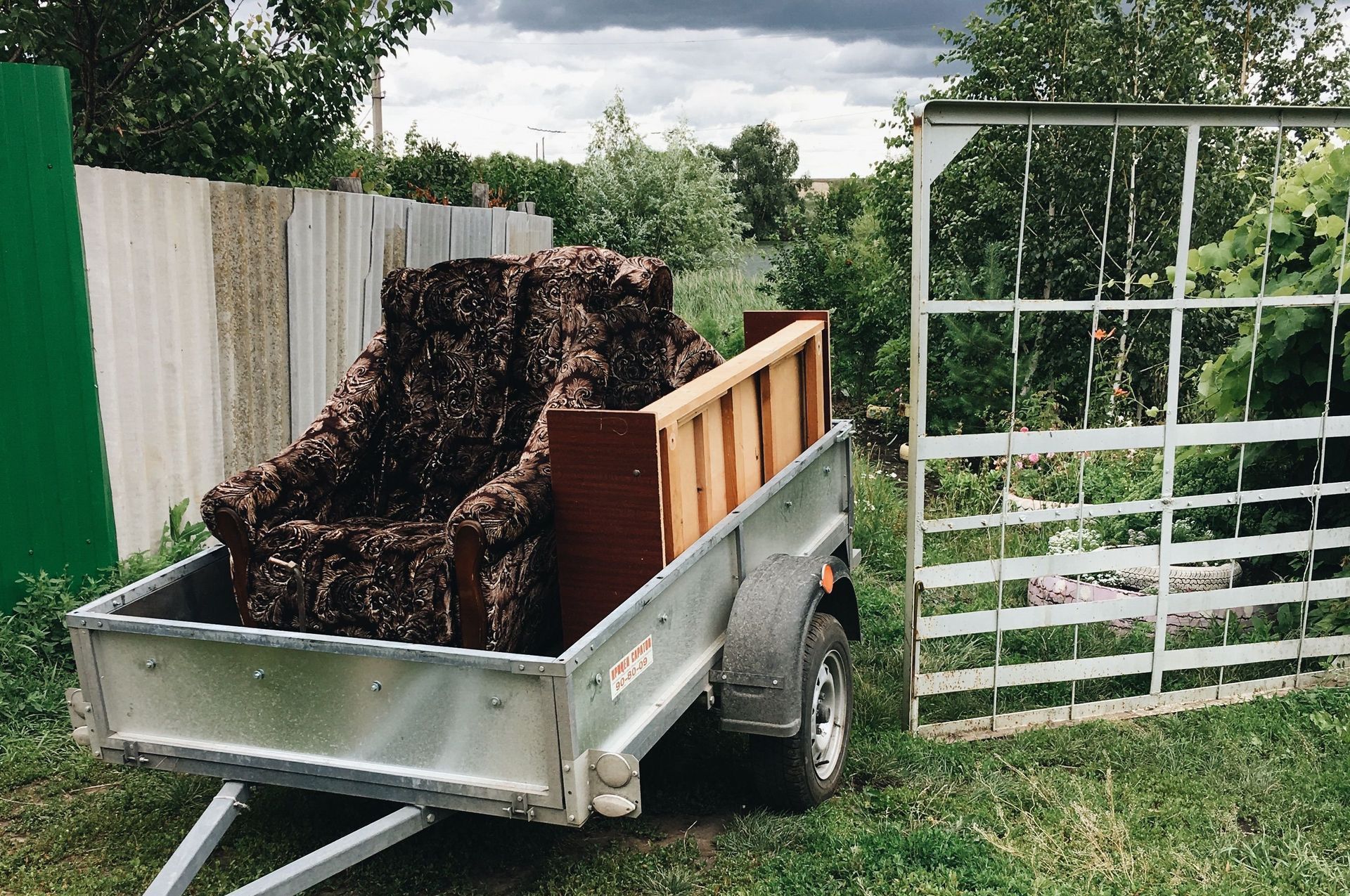 A rural setting with a trailer, vintage armchair, and a pug surrounded by greenery.