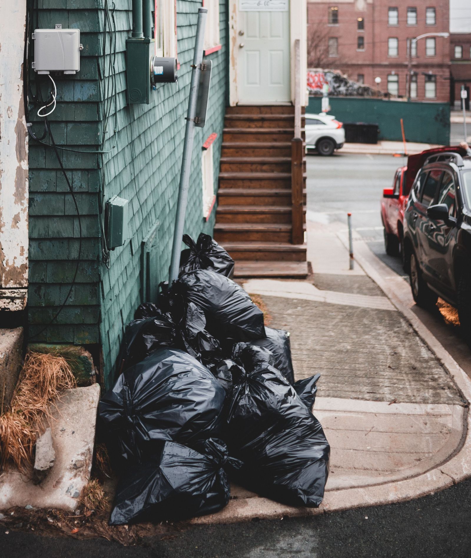 Black plastic garbage bags placed on sidewalk near colorful residential house with shabby stairs on street with buildings and cars